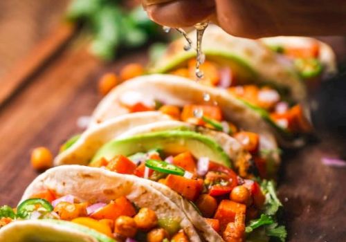 The image shows a hand squeezing lime juice over a row of vibrant, vegetable-filled tacos on a wooden surface, garnished with fresh herbs and slices of lime.