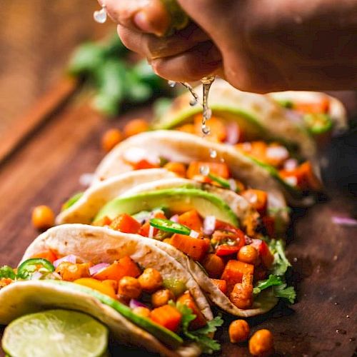 The image shows a hand squeezing lime juice over a row of vibrant, vegetable-filled tacos on a wooden surface, garnished with fresh herbs and slices of lime.