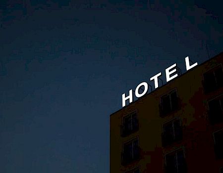 The image shows the top corner of a building at night with an illuminated "HOTEL" sign, where the letter "L" appears to be slightly tilted.