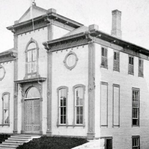 An old black-and-white photo of a two-story building with arched windows, a central entrance, and a flagpole on the roof ending the sentence.