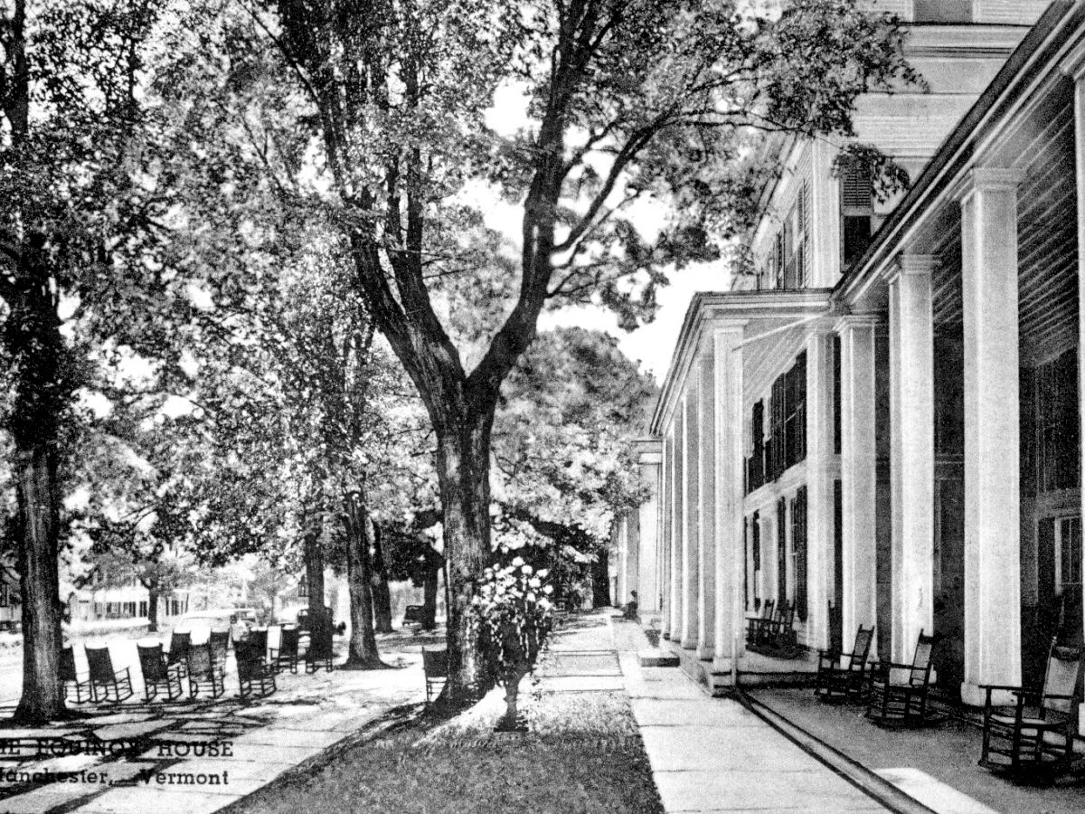 This image shows a large building with tall pillars, trees, and a shaded walkway with rocking chairs lined up in front.