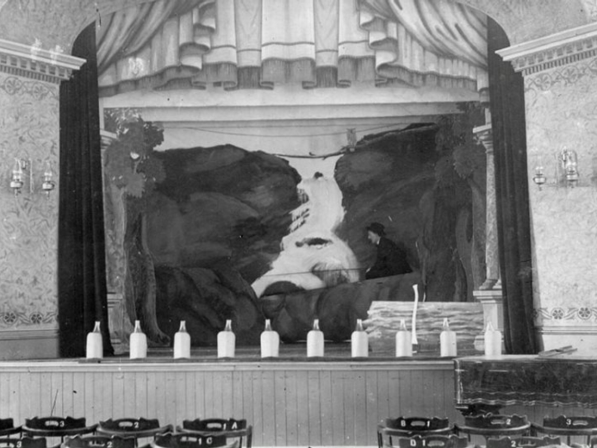The image shows a stage with a painted waterfall backdrop, curtains, chairs, and several bottles lined up on the edge of the stage.