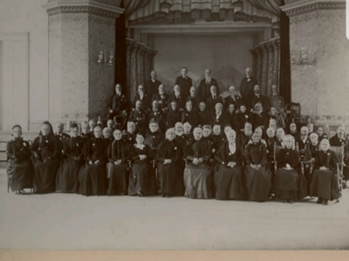 A large group of older adults is seated on stage at the Old Folks Party, Music Hall, Manchester, Vermont, dated October 24th, 1895.