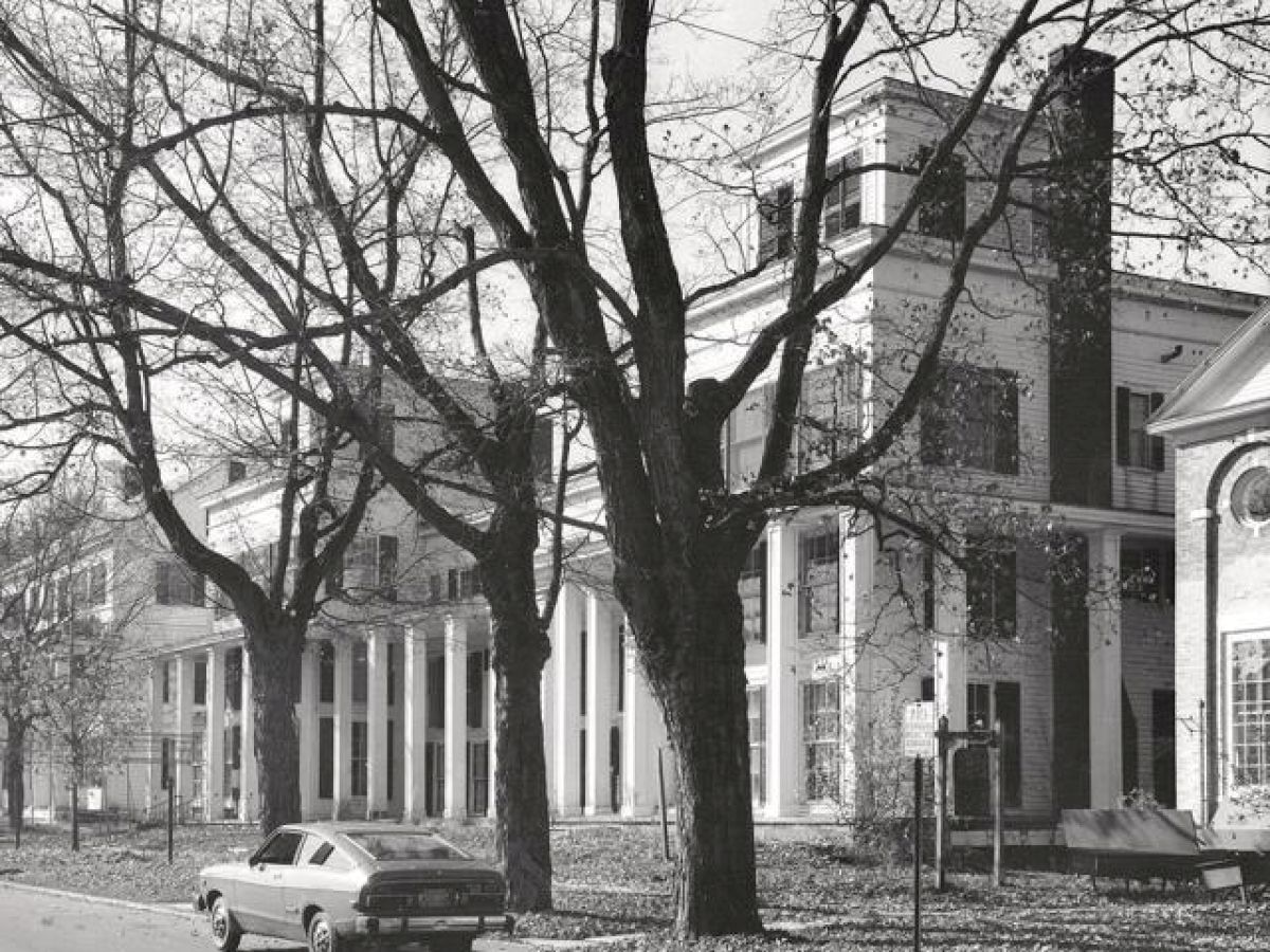 An old street scene with large trees, colonial-style buildings featuring columns, and a vintage car parked on the side of the road.