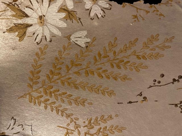 A decorative pattern shows gold fern fronds and white flowers on a muted, metallic background with subtle leaf outlines, like elegant wallpaper.