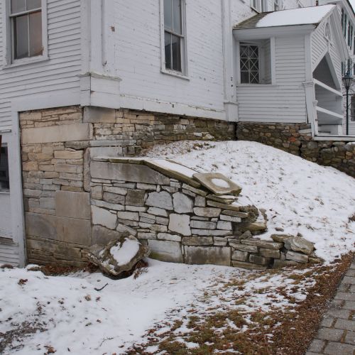 A snowy courtyard beside a white historic building, with a stone foundation wall, scattered snow patches, and a wet brick walkway.