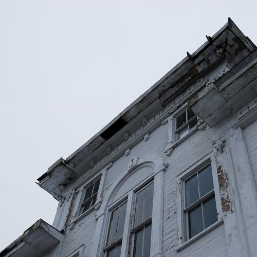 An upward view of a weathered, white wooden building with peeling paint and broken windows under an overcast sky.