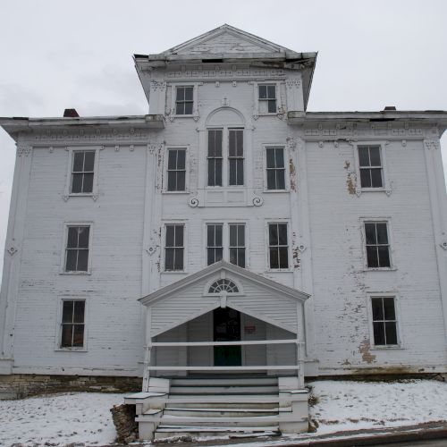 A worn white historic building with gabled entrance, many windows, brick chimneys, and snow-covered ground.