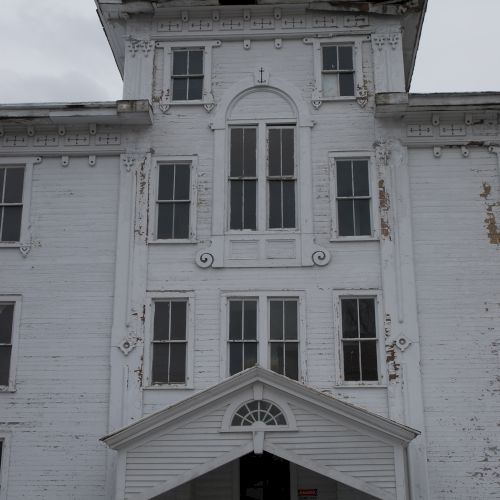 A weathered white historic building facade with arched windows, decorative trim, and a peaked gable under a gray sky.