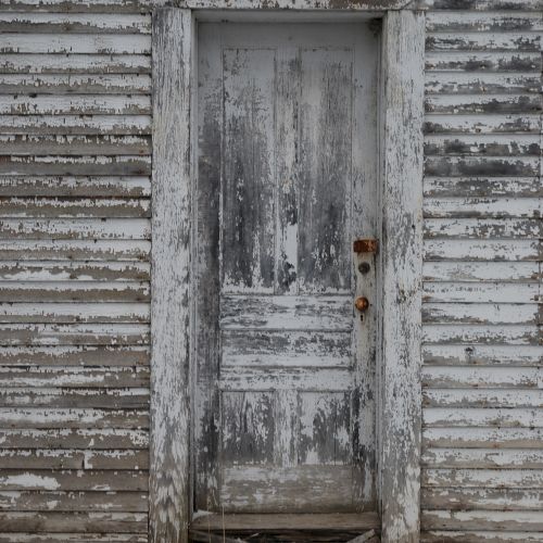 A weathered wooden door with peeling paint sits in a rusted, gray shiplap wall.