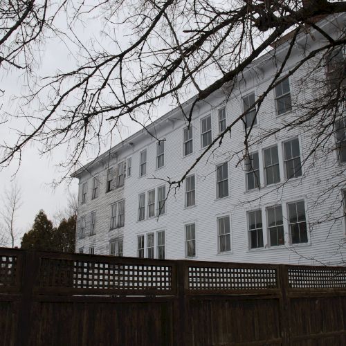 A white multi-story building sits behind a fence as leafless branches stretch across a gray sky, like winter.